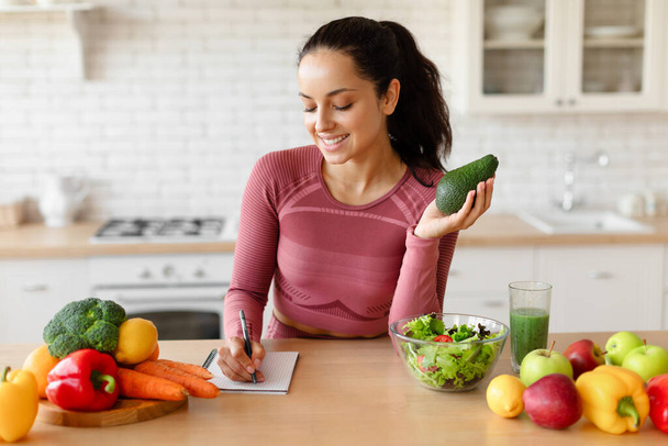 План диеты по снижению веса. Улыбающаяся фитнес-женщина записывает здоровые рецепты в свой блокнот, проведение авокадо, планирование ежедневной еды за кухонным столом со свежими овощами и фруктами в помещении. Питание - Фото, изображение