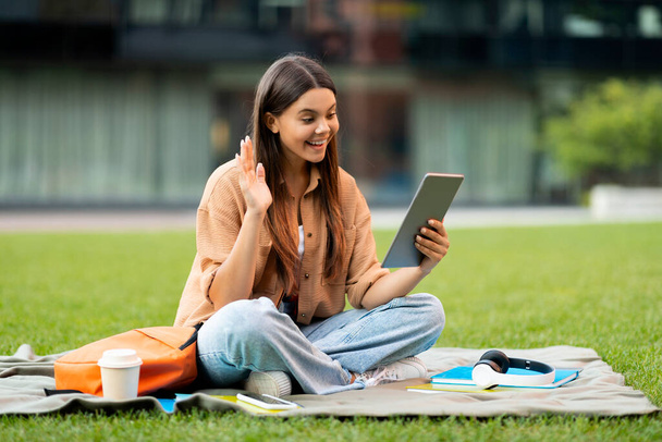 Veselá hezká brunetka mladá žena v ležérní vysokoškolské studentky odpočívající v parku, sedí na trávě, dělá domácí úkoly, pomocí digitálního tabletu, mají videohovor s přáteli, prázdný prostor. Telekomunikace - Fotografie, Obrázek