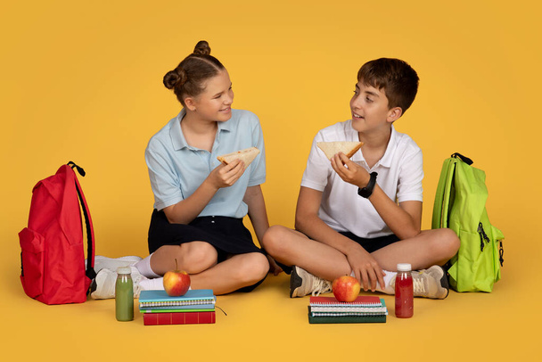 Uśmiechnięty europejski nastolatek z plecakami, jabłkami, obiadem, siedzący na podłodze z pudełkami, sokiem w szkole, odizolowany na pomarańczowym tle studia. Nauka, przyjaźń, owoce i opieka zdrowotna, przerwy żywnościowe - Zdjęcie, obraz