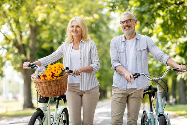 Piękna para seniorów z rowerami spacerującymi razem w słonecznym parku, - Zdjęcie, obraz