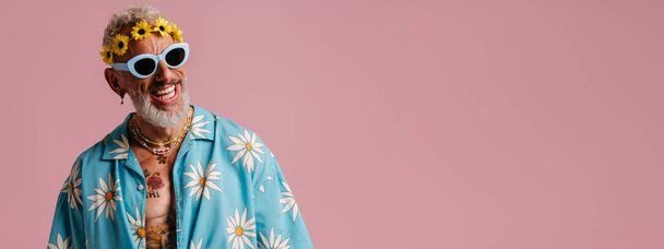 Cool senior man in stylish shirt and floral wreath on head smiling against pink background - Фото, изображение