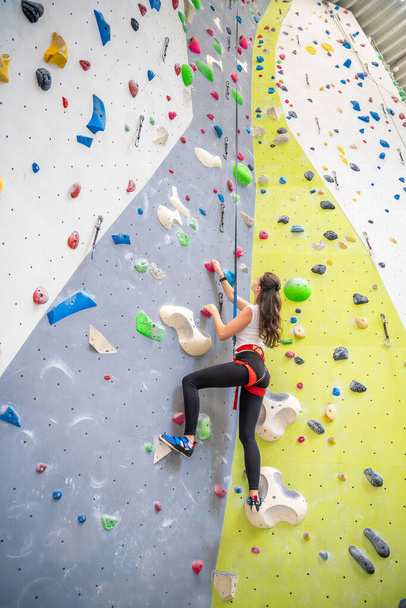 Rotsklimmer jonge vrouw opknoping op gekleurde haken op het beklimmen van kunstmatige muur binnenshuis van sport centrum van Praag. Extreme sport en bouldering concept. Hoge kwaliteit foto - Foto, afbeelding