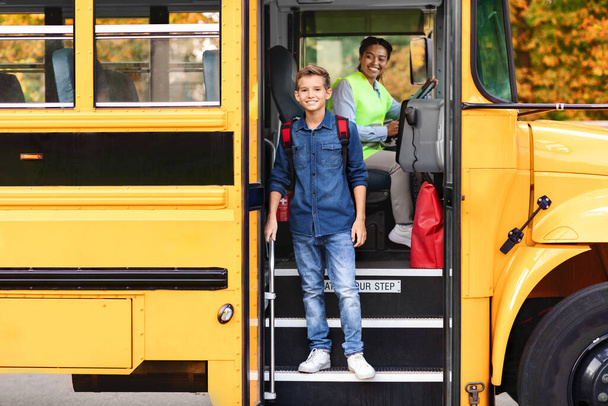 Sonriente niño preadolescente bajándose del autobús escolar amarillo, niño varón feliz bajando del vehículo cuando llega para el estudio, conductor negro con uniforme mirándolo, espacio libre - Foto, Imagen