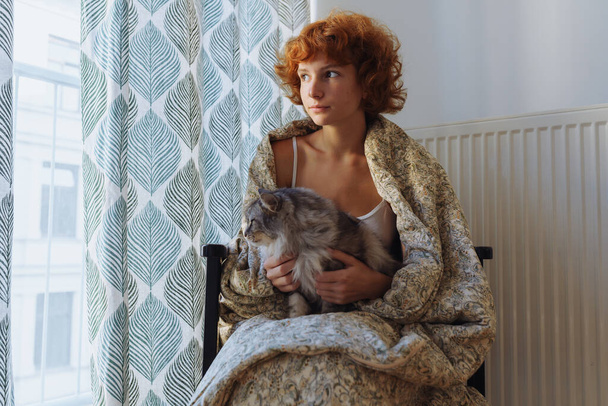 Girl hugging cat, sitting in blanket home, frozen. teen barefoot, red-haired, curly, morning, wrapped in blanket, sits hugging domestic cat, near heating radiator, large window covered with curtains. - Foto, imagen