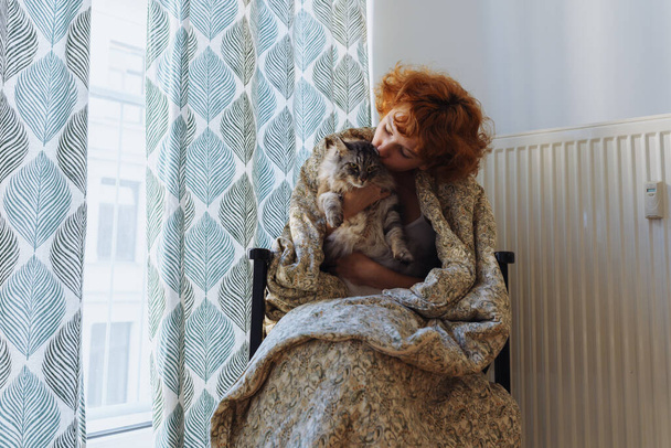 Girl hugging cat, sitting in blanket home, frozen. teen barefoot, red-haired, curly, morning, wrapped in blanket, sits hugging domestic cat, near heating radiator, large window covered with curtains. - Foto, immagini