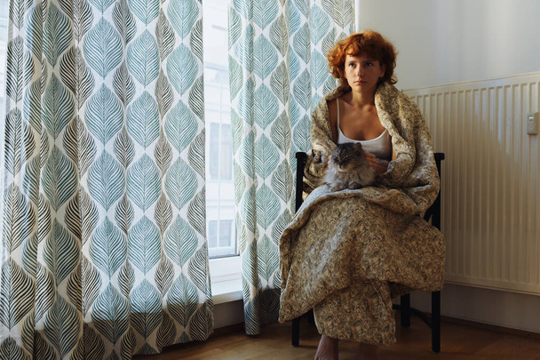 Girl hugging cat, sitting in blanket home, frozen. teen barefoot, red-haired, curly, morning, wrapped in blanket, sits hugging domestic cat, near heating radiator, large window covered with curtains. - Foto, afbeelding