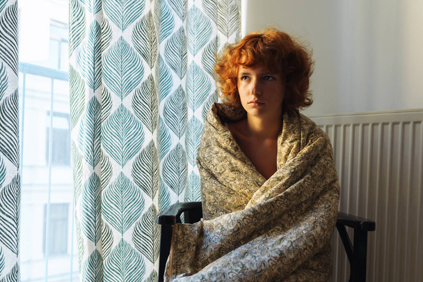 mujer joven, somnolienta, pelirroja, rizada, envuelta en manta, sentada en la silla, cerca del radiador de calefacción, congelada - Foto, Imagen