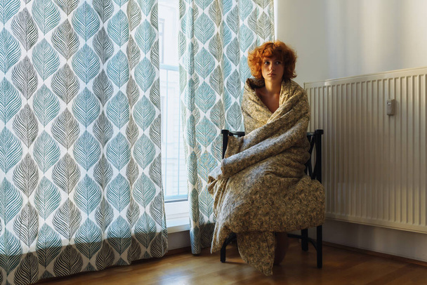 mujer joven, somnolienta, pelirroja, rizada, envuelta en manta, sentada en la silla, cerca del radiador de calefacción, congelada - Foto, imagen