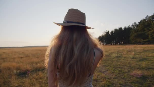 Una giovane ragazza con un cappello corre su un campo sullo sfondo di un bellissimo tramonto estivo e guarda la macchina fotografica. Al rallentatore. Primo piano - Filmati, video