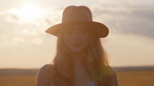 Una joven con un sombrero cruza un campo con el telón de fondo de una hermosa puesta de sol de verano y mira a la cámara. En cámara lenta. Primer plano - Imágenes, Vídeo
