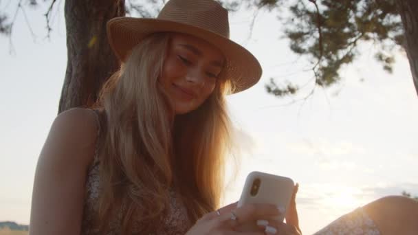Ένα νεαρό κορίτσι με καπέλο χρησιμοποιεί ένα smartphone ενώ κάθεται κάτω από ένα δέντρο με φόντο ένα όμορφο καλοκαιρινό ηλιοβασίλεμα. Αργή κίνηση. Κοντινό πλάνο - Πλάνα, βίντεο