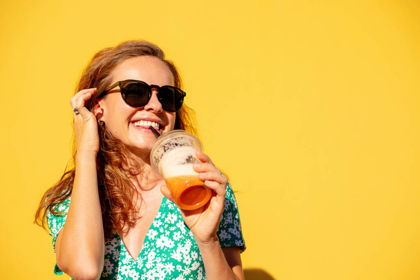 Junge selbstbewusste, unbeschwerte Frau im trendigen Sommerkleid posiert vor gelber Wand und trinkt Smoothie. - Foto, Bild