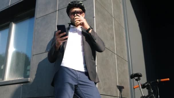 Όμορφος νεαρός άνδρας φορώντας κοστούμι σε κράνος ασφαλείας πίνοντας καφέ πριν από την οδήγηση κλασικό ποδήλατό του στη δουλειά το πρωί. - Πλάνα, βίντεο