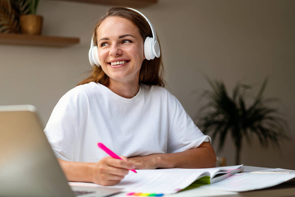 Ευτυχισμένη μαθήτρια με ακουστικά που κάθεται στο γραφείο στο σπίτι και κάνει εργασίες στο βιβλίο εργασίας. - Φωτογραφία, εικόνα