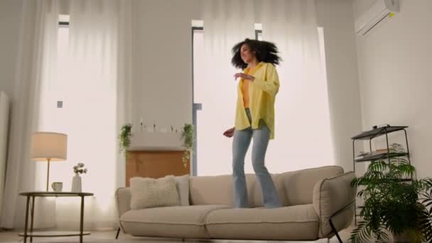 Drôle femme afro-américaine heureuse fille à la maison danse saut avoir du plaisir à tromper joie test confortable canapé doux confortable meubles pour appartement danse à la musique saut se réjouir déménagement jour - Séquence, vidéo