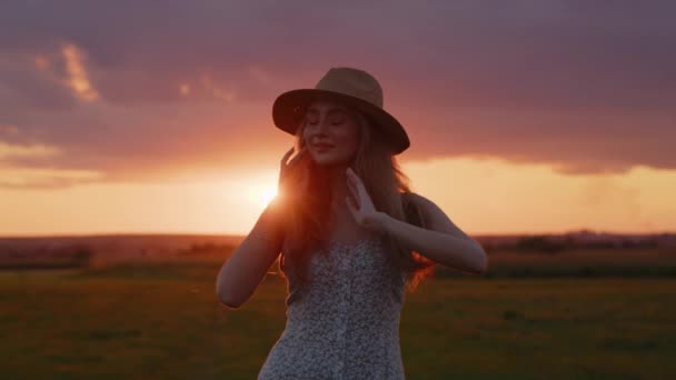 Mladá dívka v klobouku pózuje pro kameru na poli na pozadí krásného letního západu slunce a dívá se do kamery. Zpomal. Detailní záběr - Záběry, video