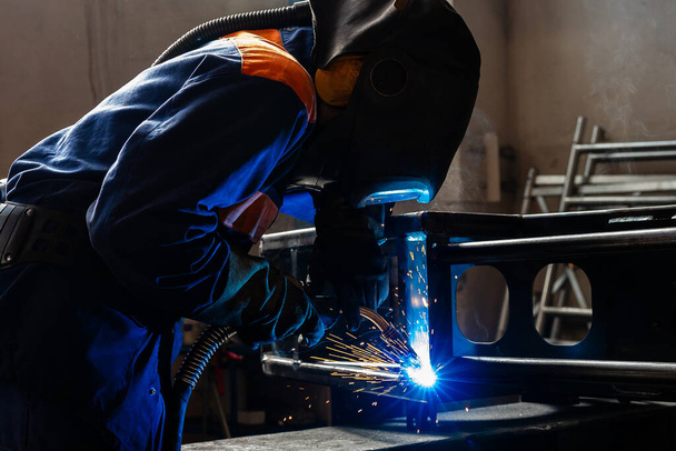 Trabajos de soldadura en una planta metalúrgica. Un soldador de casco suelda una parte metálica en una mesa de soldadura - Foto, Imagen