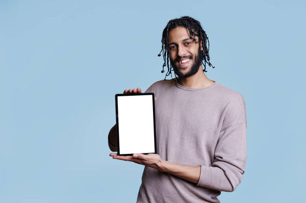 空白のタッチスクリーンでデジタルタブレットを保持し,カメラを見ている陽気なアラブ人男性. ソフトウェア広告のための空白のスクリーンの背景が付いている携帯用装置を示す笑顔の人 - 写真・画像