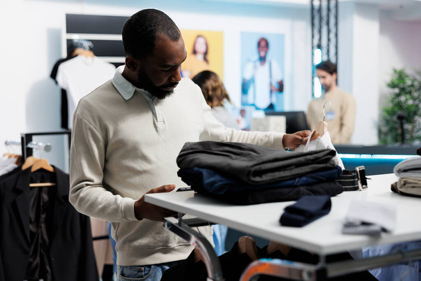 Afrikanisch-amerikanischer Mann, der in einem Einkaufszentrum Kleidung einkauft, nutzt das Smartphone, um auf die mobile Website des Bekleidungsgeschäfts zuzugreifen. Kunde wählt Hemd aus Hängegestell aus und scrollt verfügbaren Bestand in App - Foto, Bild