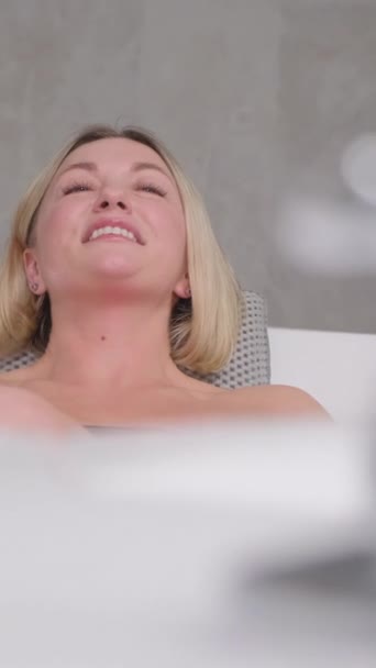 Portret uroczej kobiety siedzącej zrelaksowany w wannie z hydromasażem w luksusowym salonie spa, uśmiechnięty. Szczęśliwa kobieta leżąca w wannie, relaksująca hydroterapia w centrum urody. Pionowe wideo - Materiał filmowy, wideo