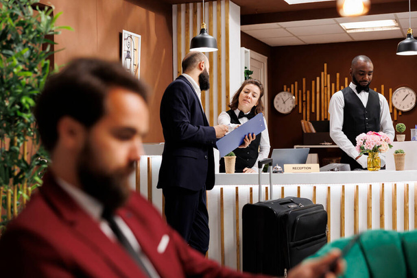 Hotel concierge assisting client at front desk, working on check in procedure to help businessman with accomodation. Traveller signing forms for booking registration, international conferences. - Foto, Imagem