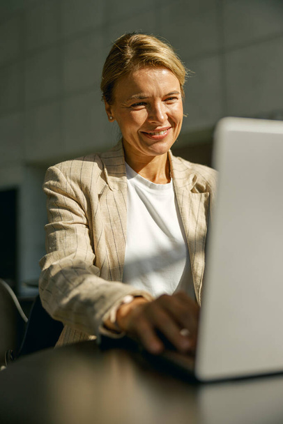 Улыбающаяся женщина-менеджер по продажам работает над ноутбуком, сидя за столом в современном коворкинге  - Фото, изображение