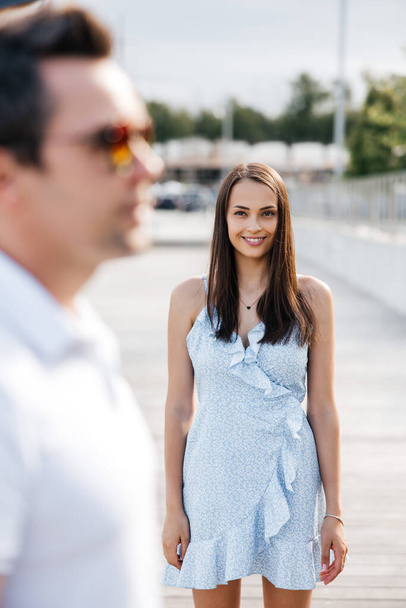 Een mooie jonge vrouw in een zomerse blauwe jurk kijkt naar haar man en glimlacht. Portret van een gelukkige vrouw die op haar man let. Verliefd stel op een wandeling - Foto, afbeelding