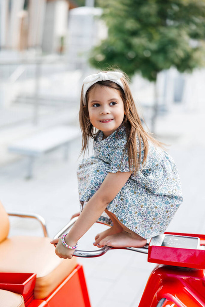 Маленька дівчинка, одягнена в літню сукню, сидить на червоній машині. Портрет усміхненої дівчинки. Щасливе і безтурботне дитинство маленької дівчинки - Фото, зображення