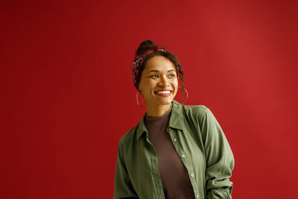 Portret uroczej kobiety odwraca wzrok z uśmiechem stojącym na czerwonym tle studia  - Zdjęcie, obraz