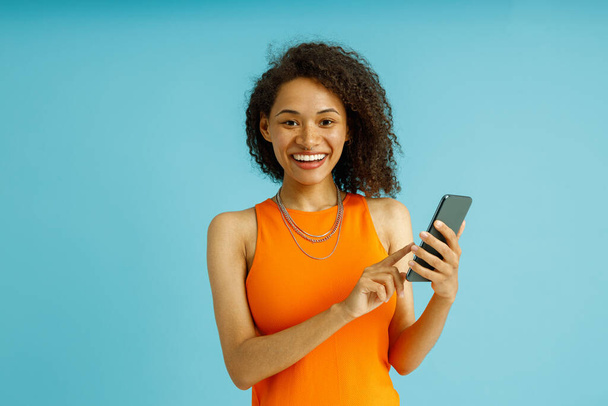 Hübsche Frau in orangefarbenem Kleid benutzt Handy, während sie auf blauem Studiohintergrund steht - Foto, Bild