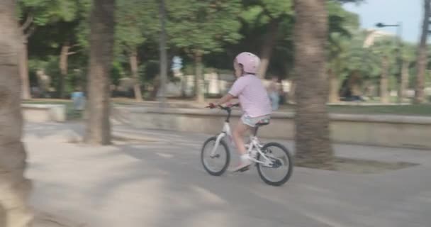 Ένα μικρό εξάχρονο καυκάσιο κορίτσι, φορώντας ένα προστατευτικό κράνος, καβαλάει ένα δίτροχο ποδήλατο κατά μήκος των μονοπατιών του πάρκου. - Πλάνα, βίντεο