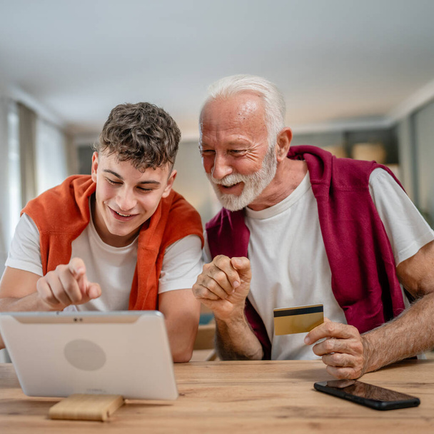 Een tiener jongeman en senior Kaukasische mannelijke grootvader zitten thuis gebruik credit card en laptop computer winkelen online gebruik internet voor online aankoop actieve moderne senior - Foto, afbeelding