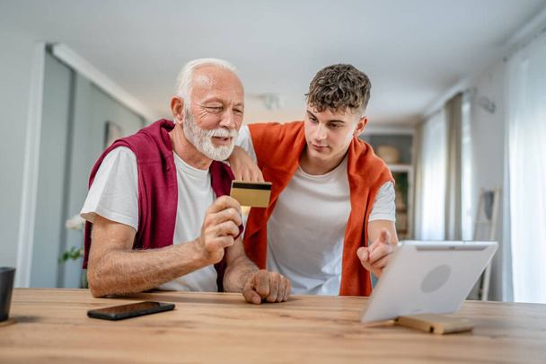 Egy tinédzser fiatalember és idősebb kaukázusi férfi nagyapja otthon ülni használja hitelkártya és laptop számítógép vásárlás online internet online vásárlás aktív modern senior - Fotó, kép