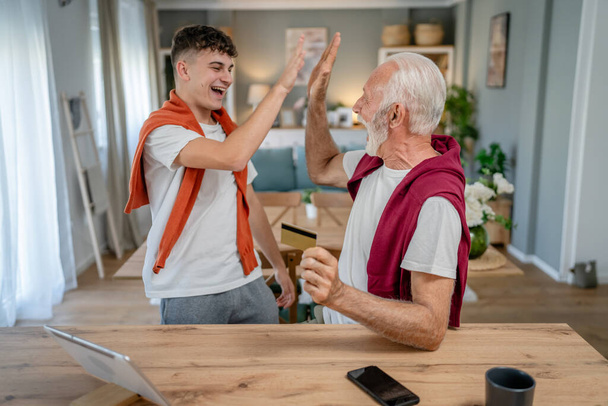Jeden nastolatek młody mężczyzna i starszy biały mężczyzna dziadek siedzieć w domu używać karty kredytowej i laptopa zakupy online korzystać z Internetu do zakupu online aktywny nowoczesny senior - Zdjęcie, obraz