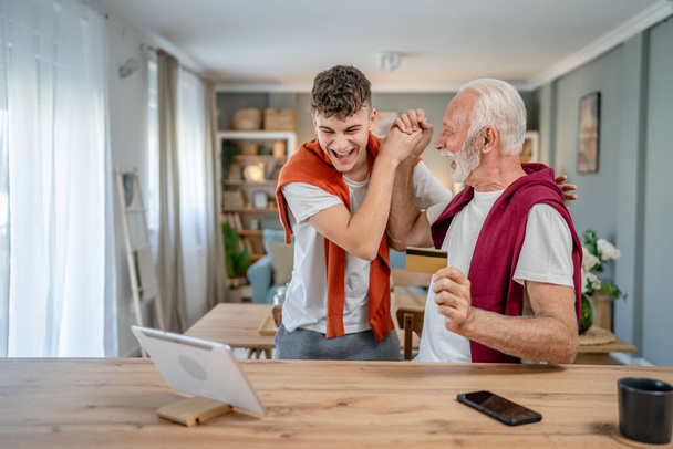 One teenager young man and senior caucasian male grandfather sit at home use credit card and laptop computer shopping online use internet for online purchase active modern senior - Photo, Image