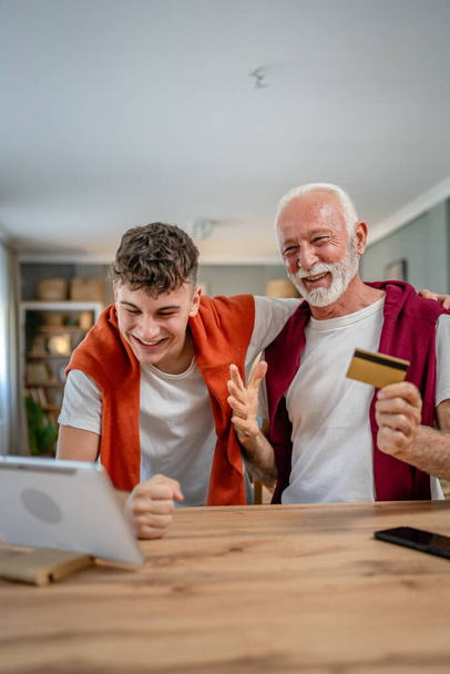 Один подросток молодой человек и старший кавказский мужчина дедушка сидеть дома использовать кредитные карты и ноутбук компьютер покупки в Интернете использовать Интернет для онлайн покупки активных современных пожилых - Фото, изображение
