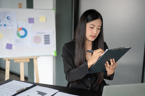 Eine professionelle asiatische Geschäftsfrau aus dem Millennium in einem formellen schwarzen Anzug konzentriert sich auf die Prüfung von Geschäftsberichten, während sie an ihrem Schreibtisch im Büro arbeitet. - Foto, Bild