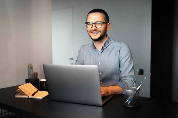Genç mutlu adamın portresi dizüstü bilgisayarda çalışıyor, gözlük ve gömlek takıyor, masada oturuyor, arka planda gri bir duvar var.. - Fotoğraf, Görsel