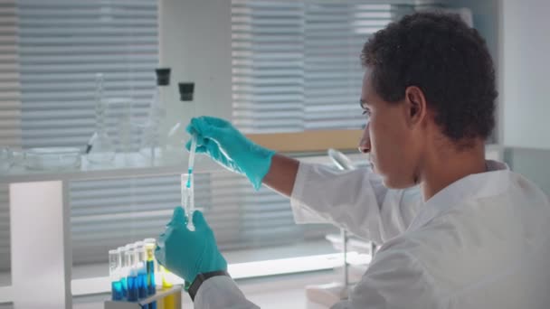 Фокусований молодий біотехнолог-біотехнолог у стерильних рукавичках та білому лабораторному пальто проводить експеримент в лабораторії, змішуючи рідини в скляній пробірці з піпеткою - Кадри, відео