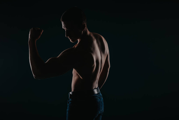 Hombre en forma mostrando bíceps. Hombre fuerte mirando sus bíceps mientras posaba con la espalda encendida - Foto, Imagen