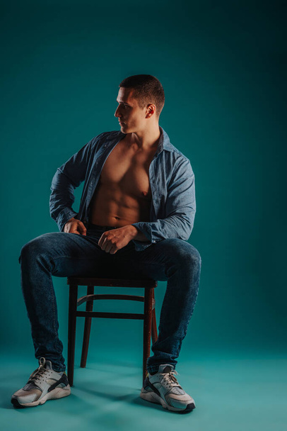 Fit homme torse nu avec chemise déboutonnée assis sur une chaise posant en studio sur un fond turquoise. Il regarde de côté. - Photo, image