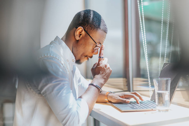Μαύρος άνδρας επιχειρηματίας που εργάζεται σε φορητό υπολογιστή κάθεται στο γραφείο δίπλα στο παράθυρο στο γραφείο. - Φωτογραφία, εικόνα