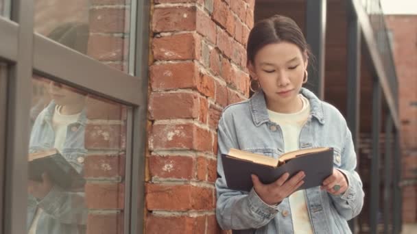 Střední pomalý portrét chytré ženy asijské vysoké školy student čtení knihy pak s úsměvem na kameru stojící mimo kampus v zamračený den - Záběry, video