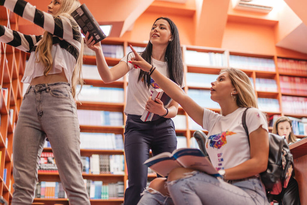 Üç genç üniversite öğrencisi kitapları kütüphanedeki kitaplığa koyuyor. - Fotoğraf, Görsel