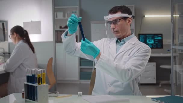 Deréktól felfelé profi kaukázusi férfi mikrobiológus védőpajzzsal és fehér köpennyel, folyadéktenyészetet vizsgálva üvegcsőben, tudományos laborban asszisztenssel - Felvétel, videó