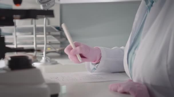 Tilt up shot of young concentrated Caucasian female scientist in eyeglasses and white lab coat using microscope while doing research in modern scientific laboratory - Imágenes, Vídeo