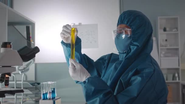 Πλάγια όψη μέση προς τα πάνω πλάνο του άνδρα βιοχημικός σε προστατευτική κάλυψη ομοιόμορφη εξέταση υγρών σε γυάλινους δοκιμαστικούς σωλήνες, ενώ κάνει έρευνα στο εργαστήριο - Πλάνα, βίντεο