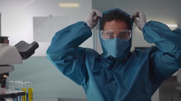 現代の研究室で働く彼の保護カバーの顔のマスクとフードを取っている間,カメラを見ている若いBiracial男性科学者の肖像画を待って下さい - 映像、動画