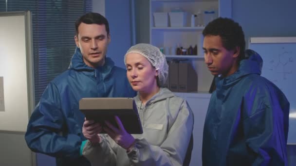Tiro medio de equipo multiétnico de científicos con batas protectoras usando tableta digital mientras investigan juntos en un laboratorio moderno - Metraje, vídeo
