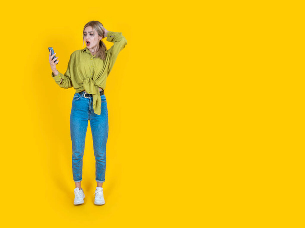 Confused shocked young woman, full body front view beautiful blonde confused shocked young woman. Puzzled stressed sad caucasian lady holding using mobile cell phone isolated on yellow background. - Photo, Image